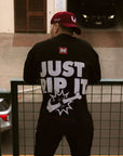 Back view of a person wearing a black BJJ t-shirt with the bold "JUST RIP IT" graphic, leaning on a railing. The red and white brand logo is visible near the collar.