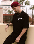 Man wearing an oversized black BJJ t-shirt with a front chest logo and a maroon baseball cap. He is leaning against a wall on an outdoor balcony in a casual Jiu-Jitsu lifestyle shot.