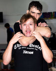Two individuals in a grappling pose with one person on top, wearing a black shirt with text.