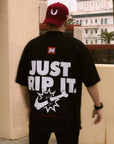Back view of a man wearing a maroon cap and an oversized black BJJ t-shirt with a large "JUST RIP IT" graphic, promoting a strong grappling or Jiu-Jitsu lifestyle.