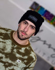 Person wearing a black beanie with 'NATION' logo and a patterned sweater, standing against a colorful abstract background.