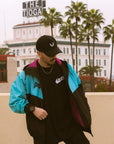 Model in a black, teal, and magenta BJJ windbreaker hoodie jacket over a logo t-shirt, standing on a rooftop with the historic 'THE TIOGA' sign and palm trees in the background.