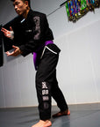 A martial artist in a black Brazilian Jiu-Jitsu uniform with a purple belt, posing on a gray mat in an indoor training room.