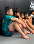Young boy wearing multicolor One For All BJJ Kids Rash Guard sitting on gym mat with other kids in background