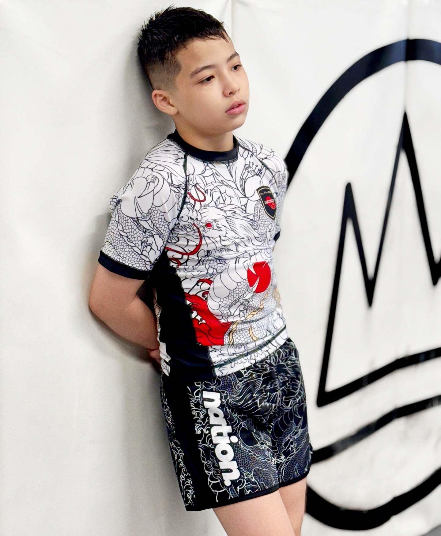 Boy wearing Rising Dragon white short sleeve Kids BJJ rash guard with black collar and dragon design leaning against wall