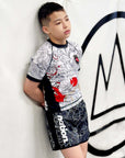Boy wearing Rising Dragon white short sleeve Kids BJJ rash guard with black collar and dragon design leaning against wall