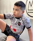 Young boy wearing black Rising Dragon Kids BJJ grappling shorts and matching rash guard sitting on mat
