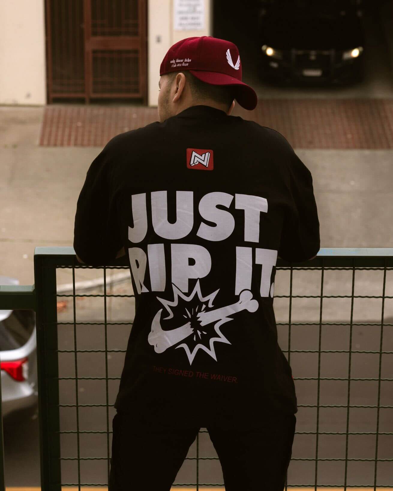 Back view of a person wearing a black BJJ shirt with the bold "JUST RIP IT" graphic, leaning on a railing. The red and white brand logo is visible near the collar.