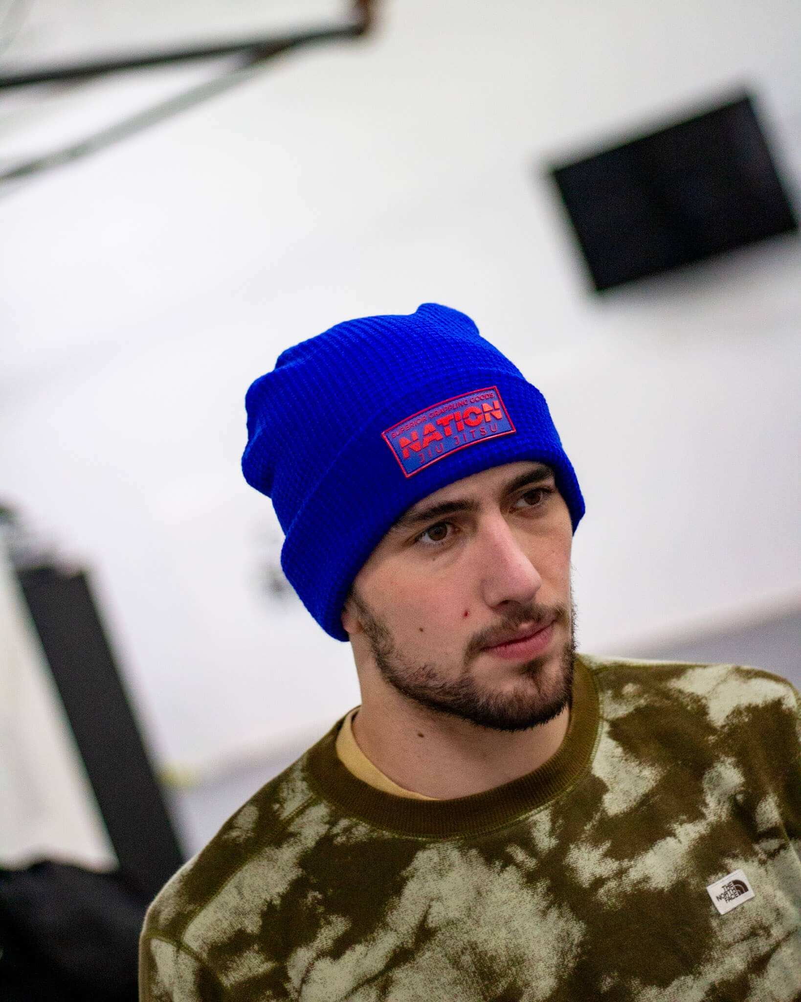 Man wearing a blue beanie with a logo, standing indoors against a white wall.