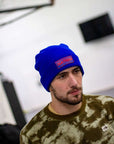 Man wearing a blue beanie with a logo, standing indoors against a white wall.