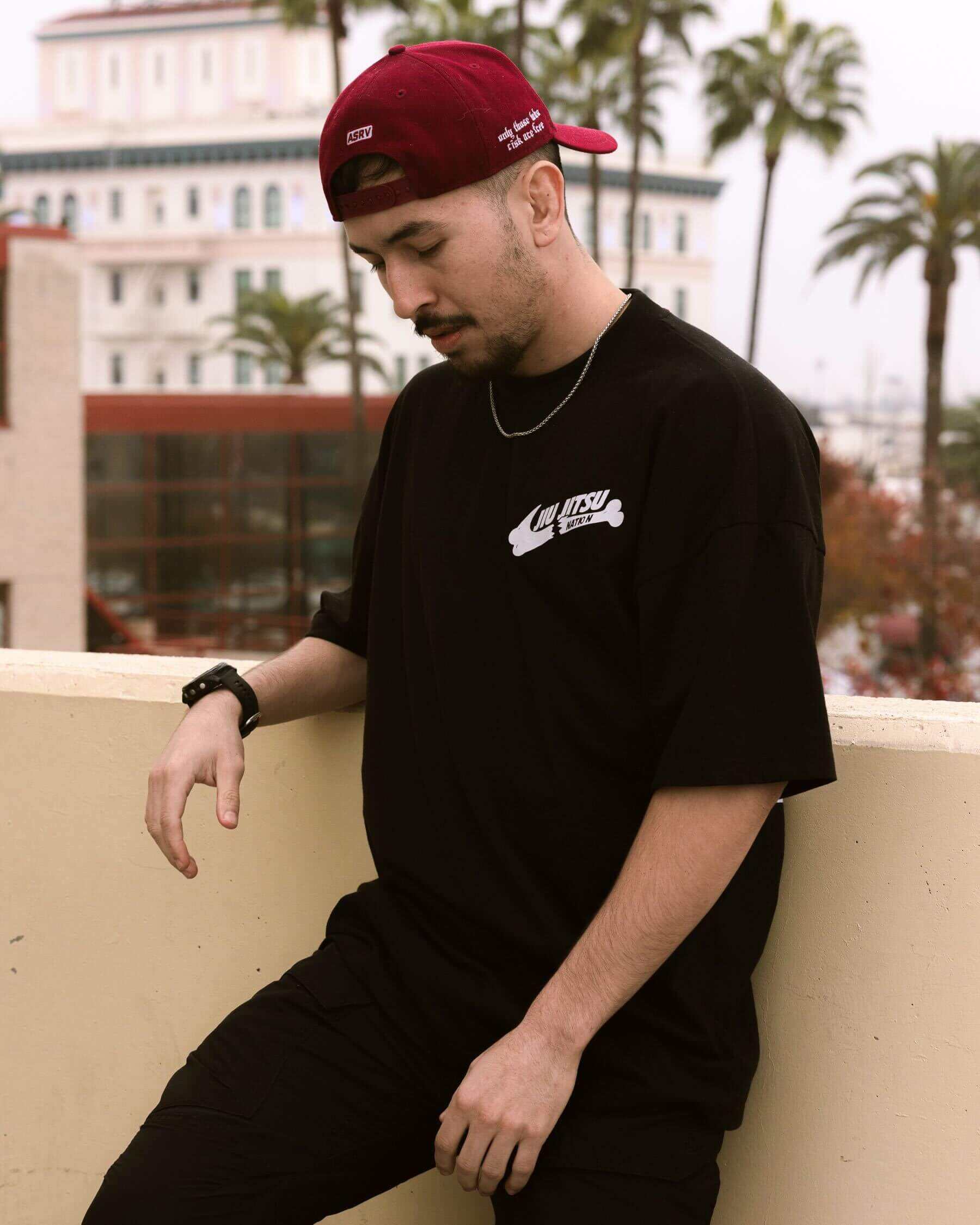 Man wearing an oversized black BJJ t-shirt with a front chest logo and a maroon baseball cap. He is leaning against a wall on an outdoor balcony in a casual Jiu-Jitsu lifestyle shot.