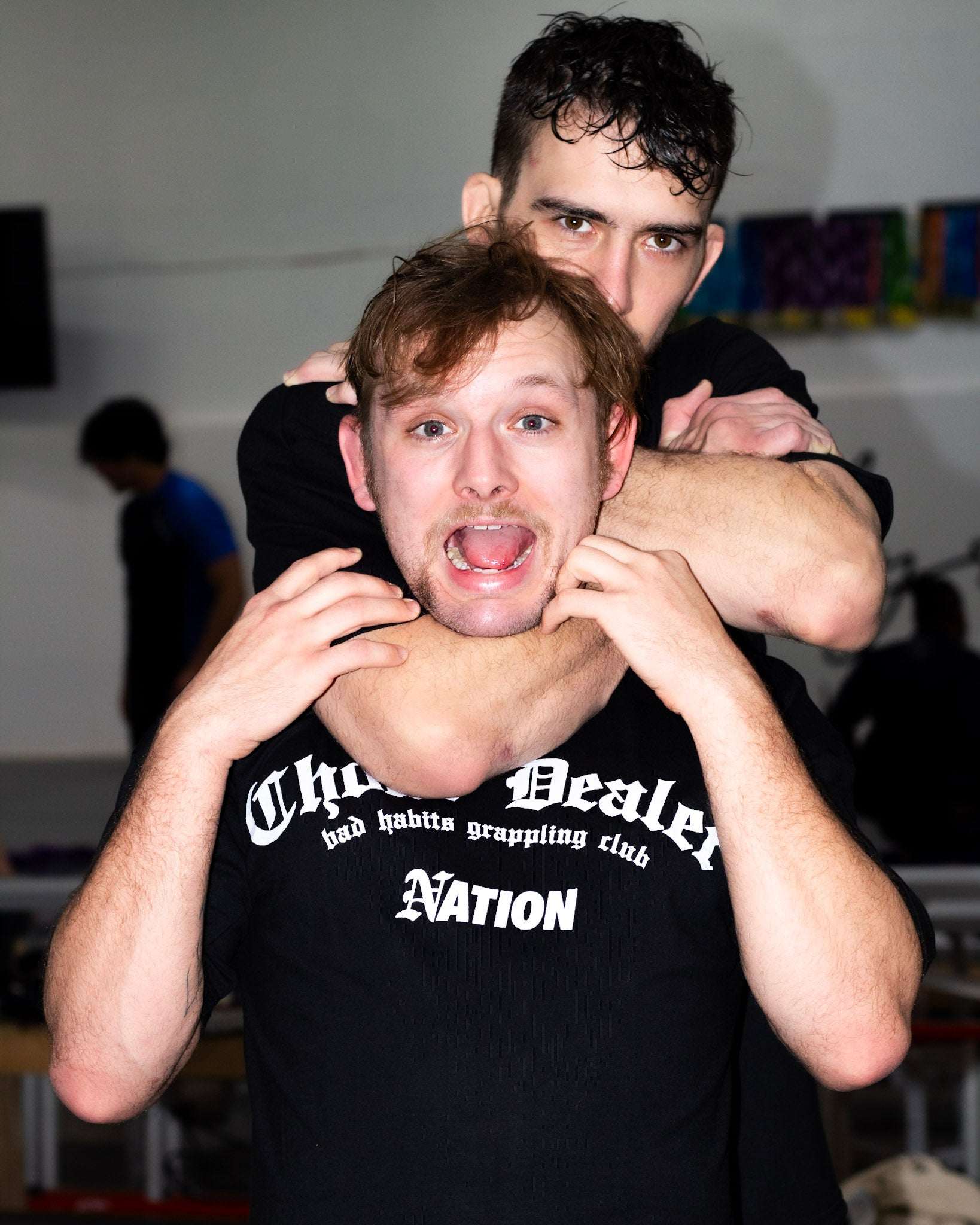 Two individuals in a grappling pose with one person on top, wearing a black shirt with text.