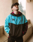 Frontal view of a man wearing the teal and black BJJ hoodie windbreaker jacket fully zipped, revealing the magenta mesh hood lining. He is also wearing a black cap.