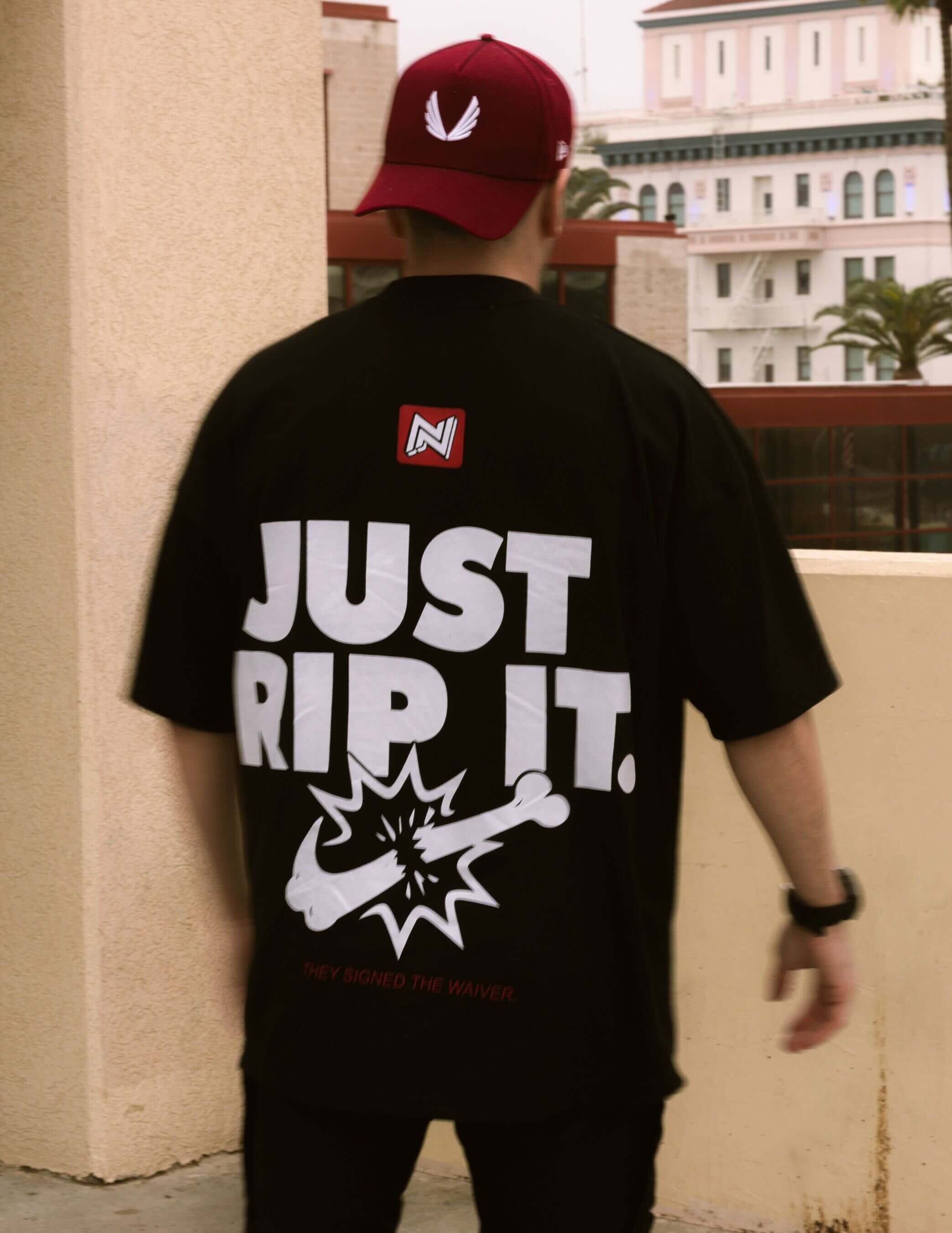 Back view of a man wearing a maroon cap and an oversized black BJJ t-shirt with a large "JUST RIP IT" graphic, promoting a strong grappling or Jiu-Jitsu lifestyle.