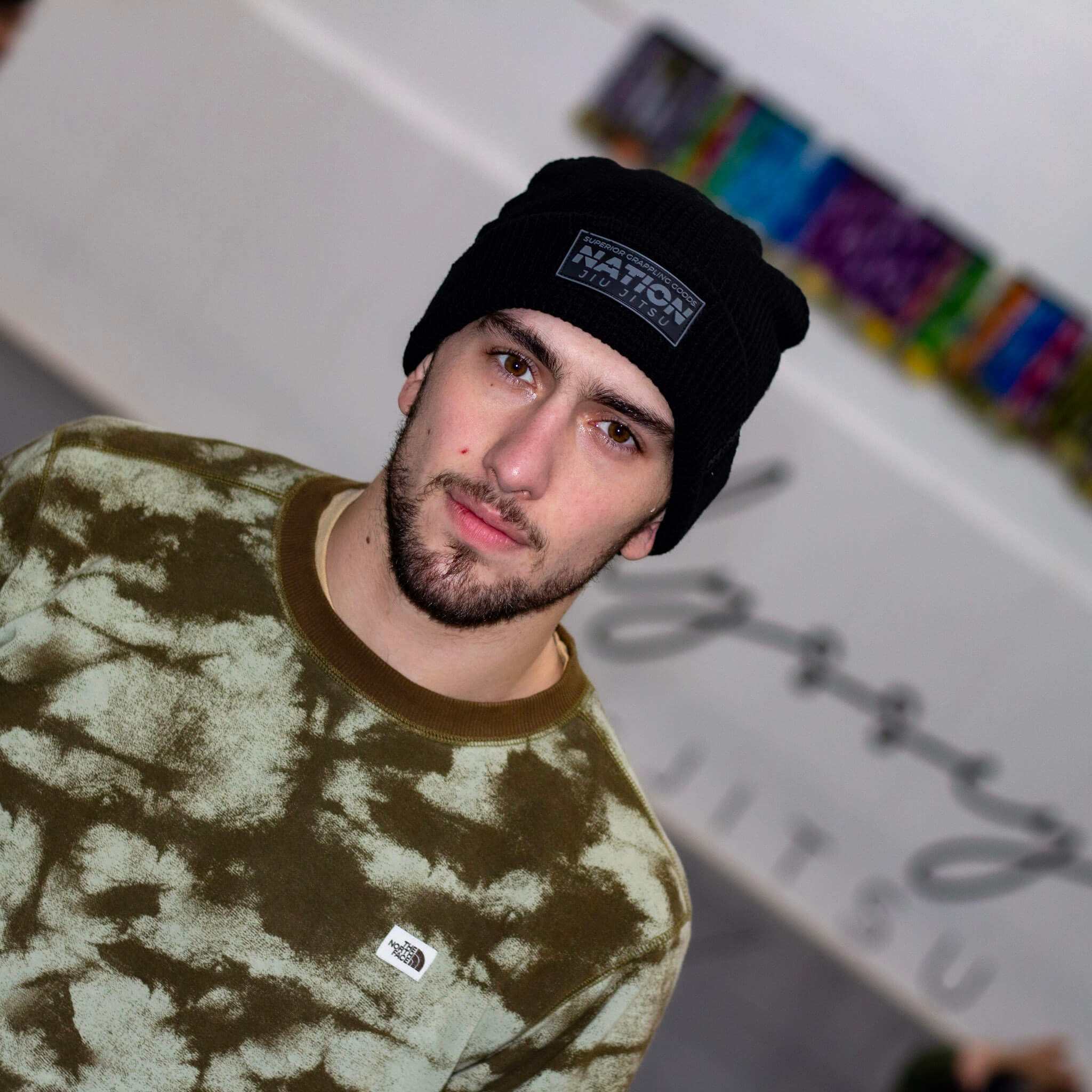 Person wearing a black beanie with &#39;NATION&#39; logo and a patterned sweater, standing against a colorful abstract background.
