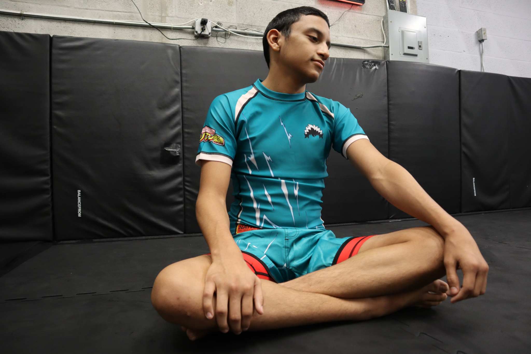 Person sitting on a mat in a martial arts or fitness studio wearing a blue athletic outfit.