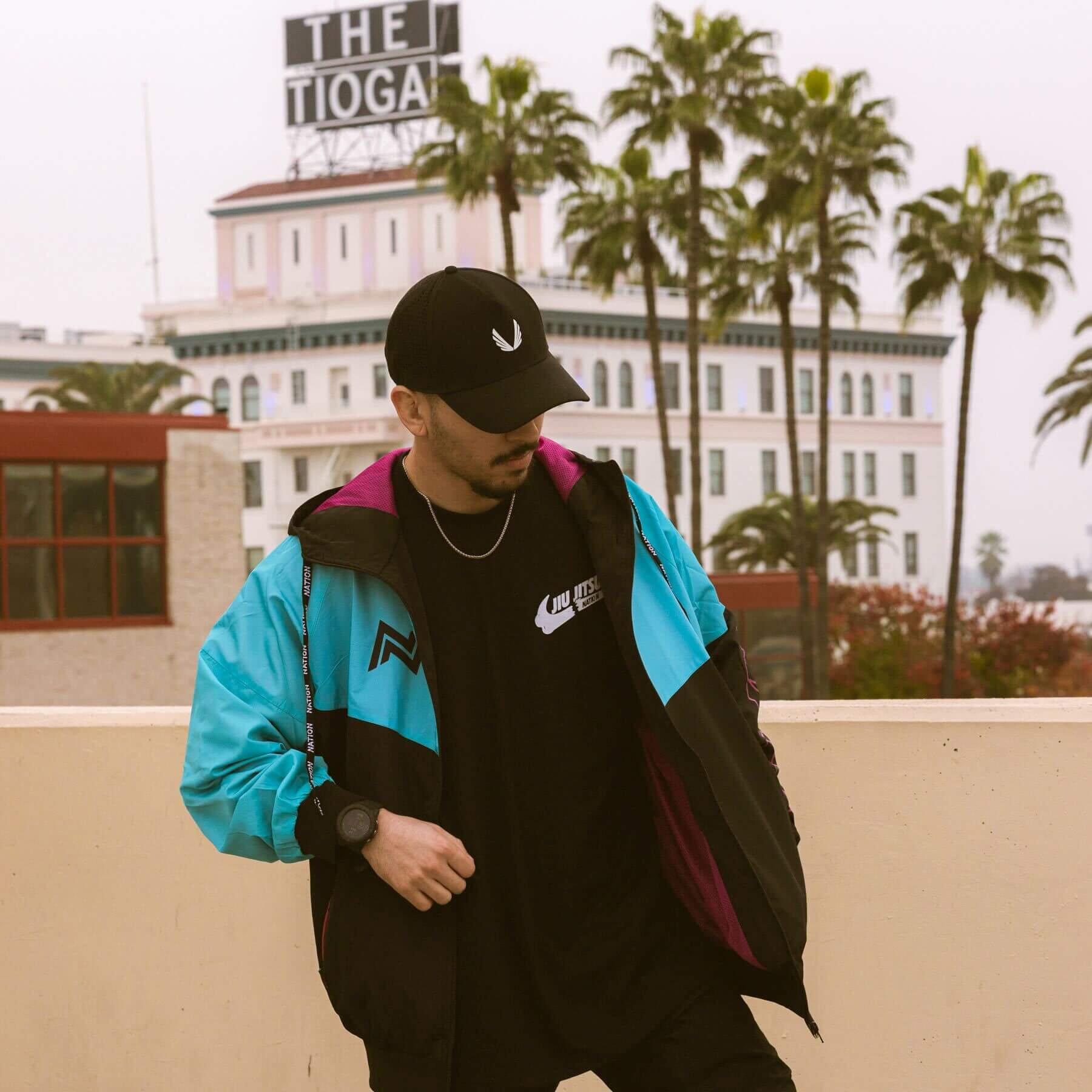 Model in a black, teal, and magenta BJJ windbreaker hoodie jacket over a logo t-shirt, standing on a rooftop with the historic &#39;THE TIOGA&#39; sign and palm trees in the background.