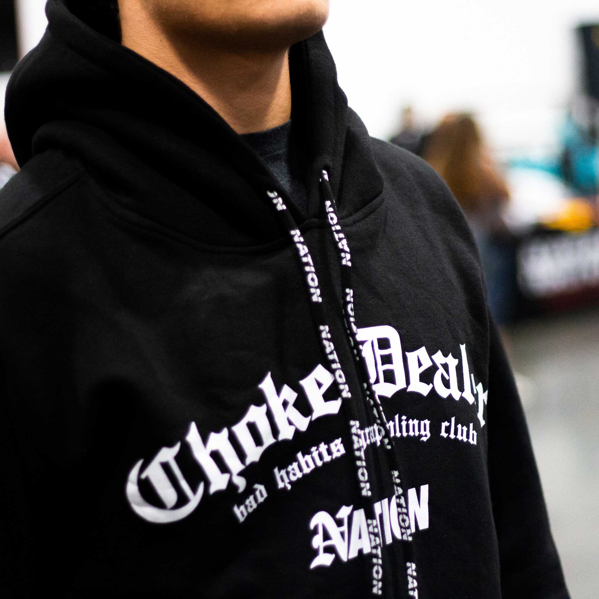 Black BJJ hoodie choke dealer with white gothic text and branding on a blurred background