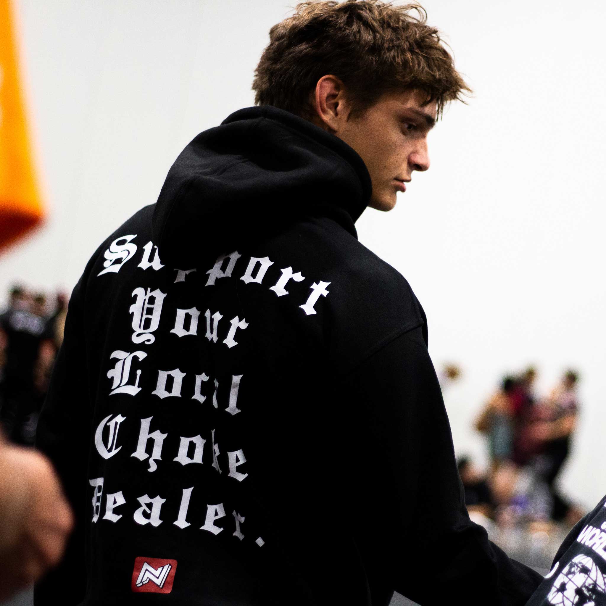Person wearing a Black BJJ hoodie choke dealer with white gothic text and branding on a blurred background
