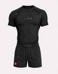 A black athletic compression shirt and matching shorts set on a light gray background. The shirt features a short-sleeve design with mesh panels on the shoulders and sides, a subtle metallic logo on the chest, and an elastic waistband.
