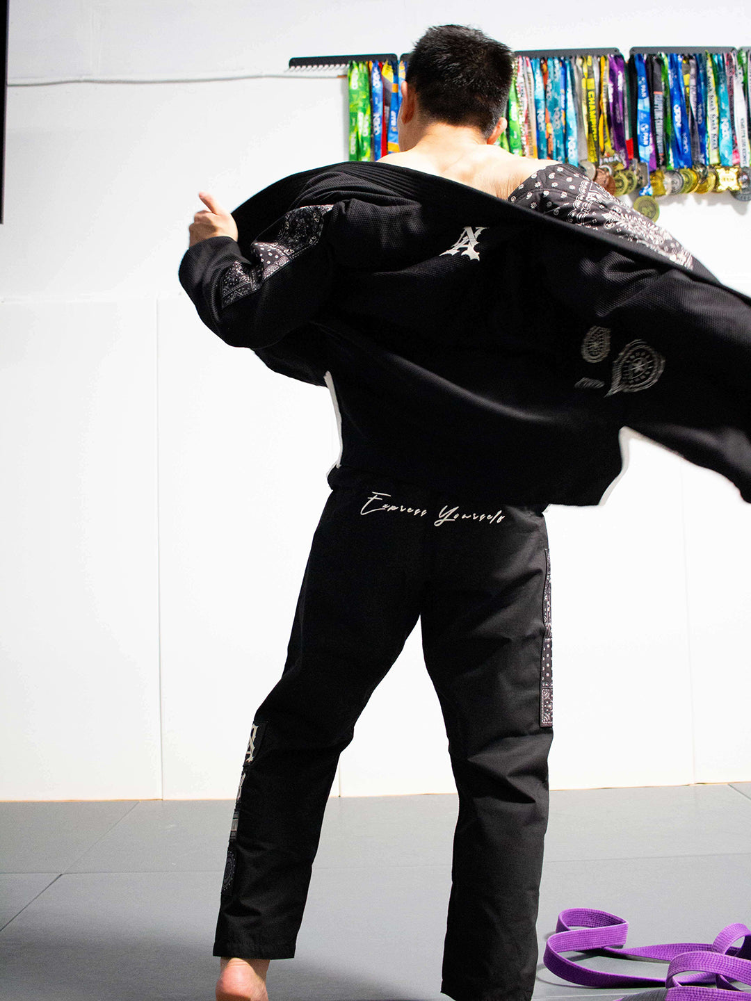 A man wearing a black martial arts gi jacket and black pants, shown from behind as he opens the jacket to reveal a patterned lining. The gi has white script embroidery on the back of the pants and a white 'A' logo on the back of the jacket.