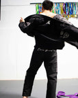 A man wearing a black martial arts gi jacket and black pants, shown from behind as he opens the jacket to reveal a patterned lining. The gi has white script embroidery on the back of the pants and a white 'A' logo on the back of the jacket.