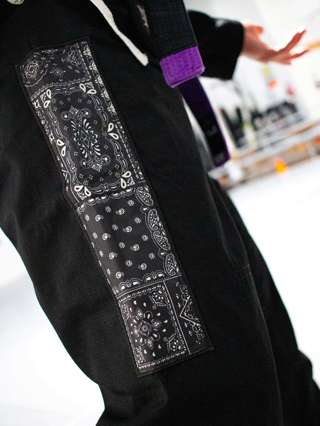 A close-up of a black Brazilian jiu jitsu kimono pants, featuring three vertical rectangular patches with white and gray bandana prints on the left side. A purple belt is visible around the wearer's waist, and a blurred indoor background is present.