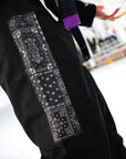 A close-up of a black Brazilian jiu jitsu kimono pants, featuring three vertical rectangular patches with white and gray bandana prints on the left side. A purple belt is visible around the wearer's waist, and a blurred indoor background is present.