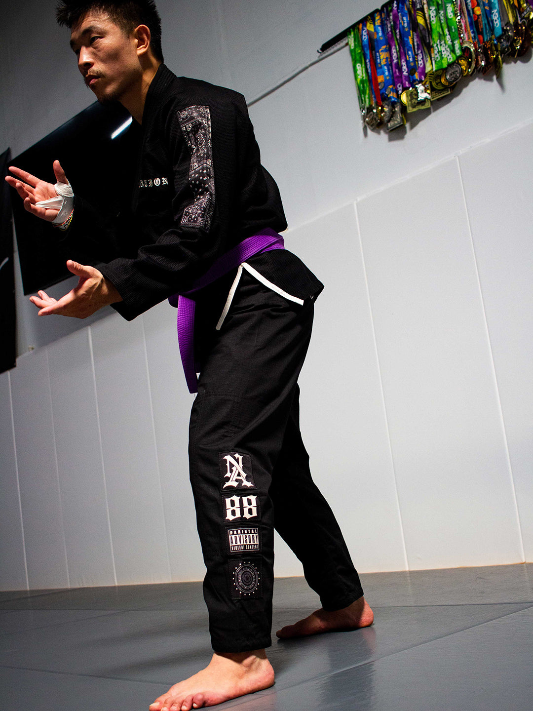 A martial artist in a black Brazilian Jiu-Jitsu uniform with a purple belt, posing on a gray mat in an indoor training room.