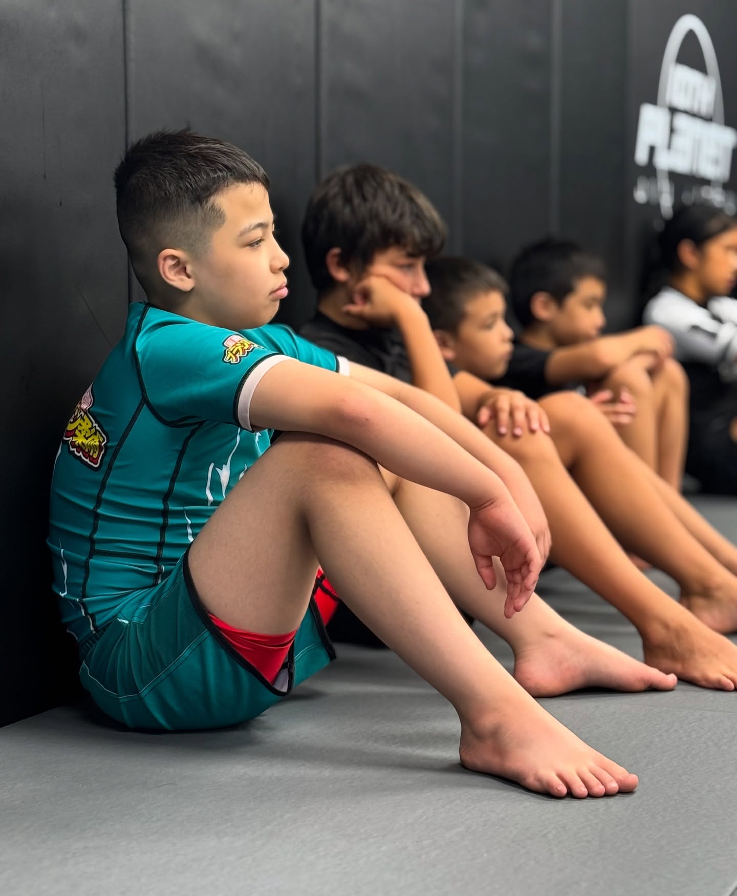 Young boy wearing multicolor One For All BJJ Kids Rash Guard sitting on gym mat with other kids in background