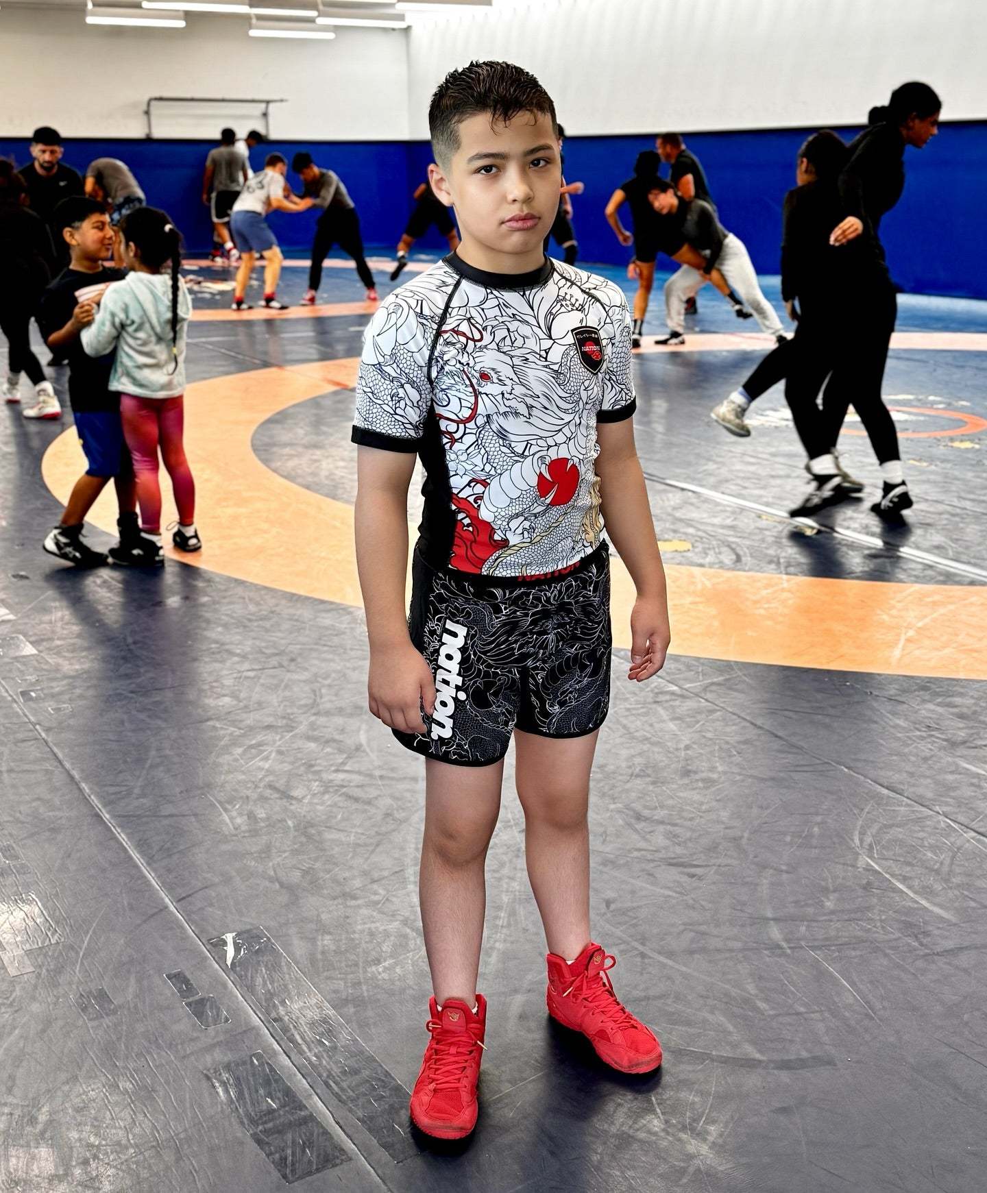 Youth wearing Rising Dragon Kids BJJ rash guard set with dragon design and black grappling shorts on wrestling mat