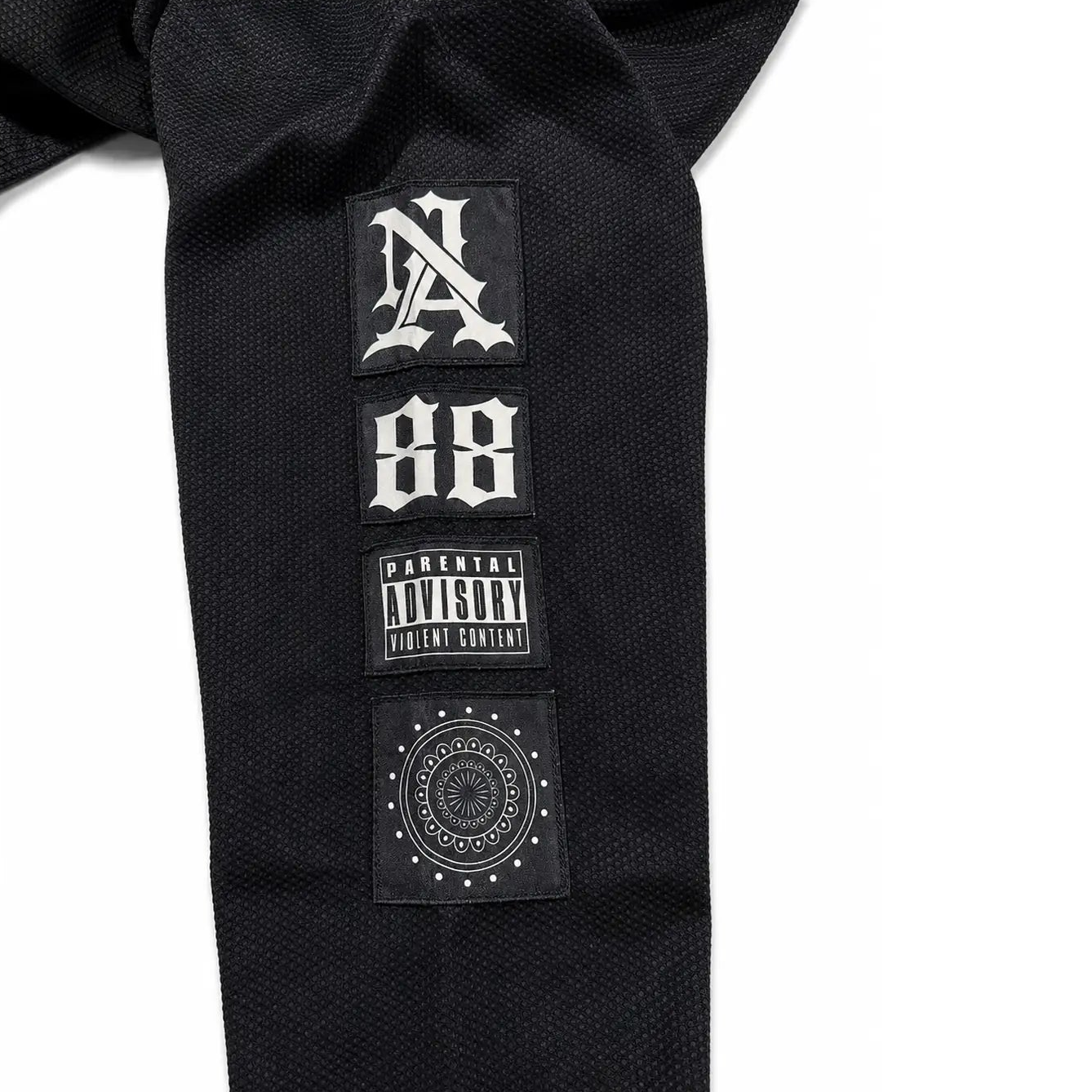 Close-up of a black textured garment sleeve featuring three rectangular patches: a stylized 'NA' patch, a '88' patch, and a 'Parental Advisory' label patch, on a white background.