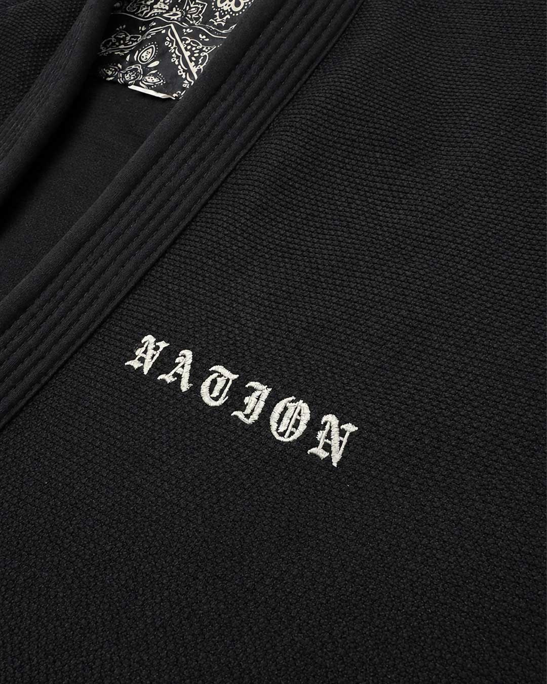 Close-up of a black textured knit Brazilian jiu jitsu kimono jacket with white embroidered 'NATION' lettering on the chest and a patterned inner neck label.