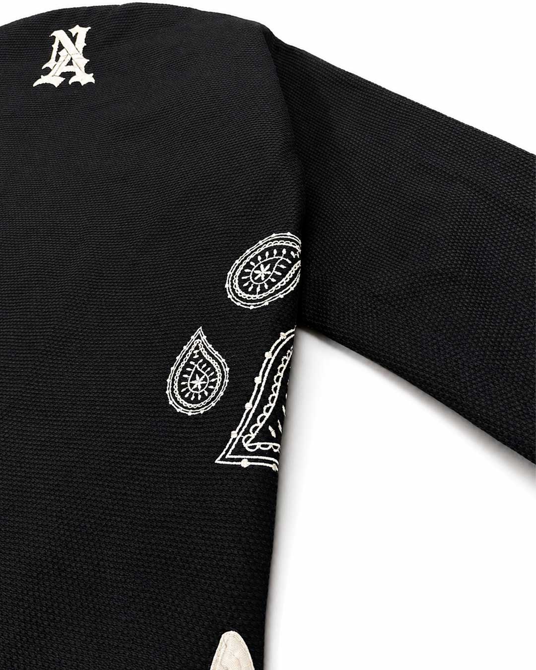 Close-up of a black Brazilian jiu jitsu kimono jacket, likely a sweater or cardigan, featuring white embroidered paisley bandana-style motifs and a white monogram letter 'A' near the shoulder.