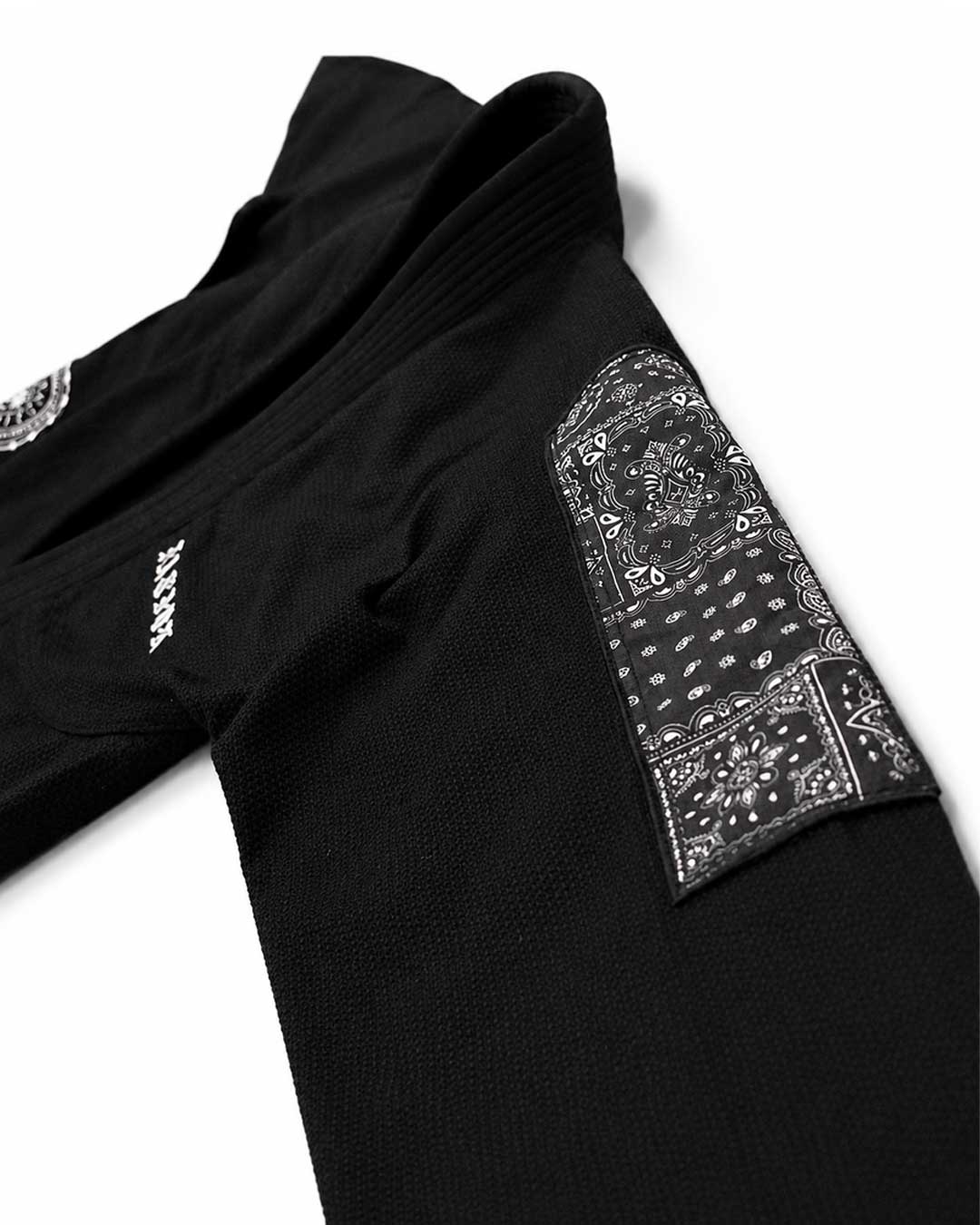 Close-up of a black jiu jitsu gi jacket laid flat on a white background, featuring a large black-and-white bandana print patch on the sleeve and small white logo text on the side.