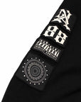 Close-up of a black garment sleeve with four rectangular embroidered patches in a vertical row. The patches are white and feature stylized lettering, a 'Parental Advisory' label, and a circular mandala-like emblem.