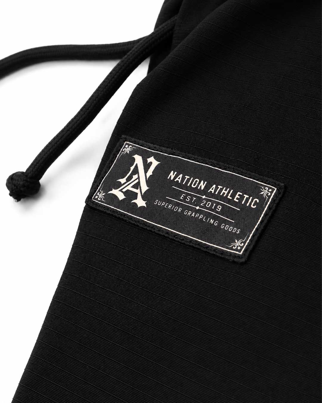 Close-up of a black ribbed fabric garment with a black drawstring and a rectangular sewn patch reading NATION ATHLET EST. 2019 SUPERIOR GRAMPLING GOODS on a white background.